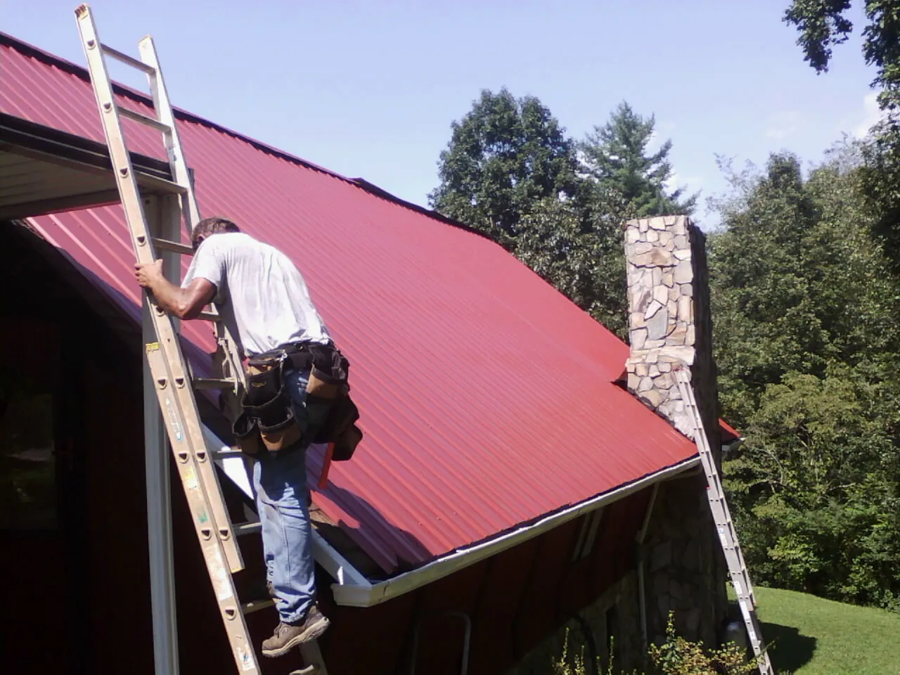 Roofer on a ladder beside a red metal roof and stone chimney in El Paso